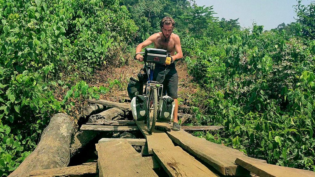 Explorer Jude Kriwald pushing his bike over a wooden bridge in Liberia